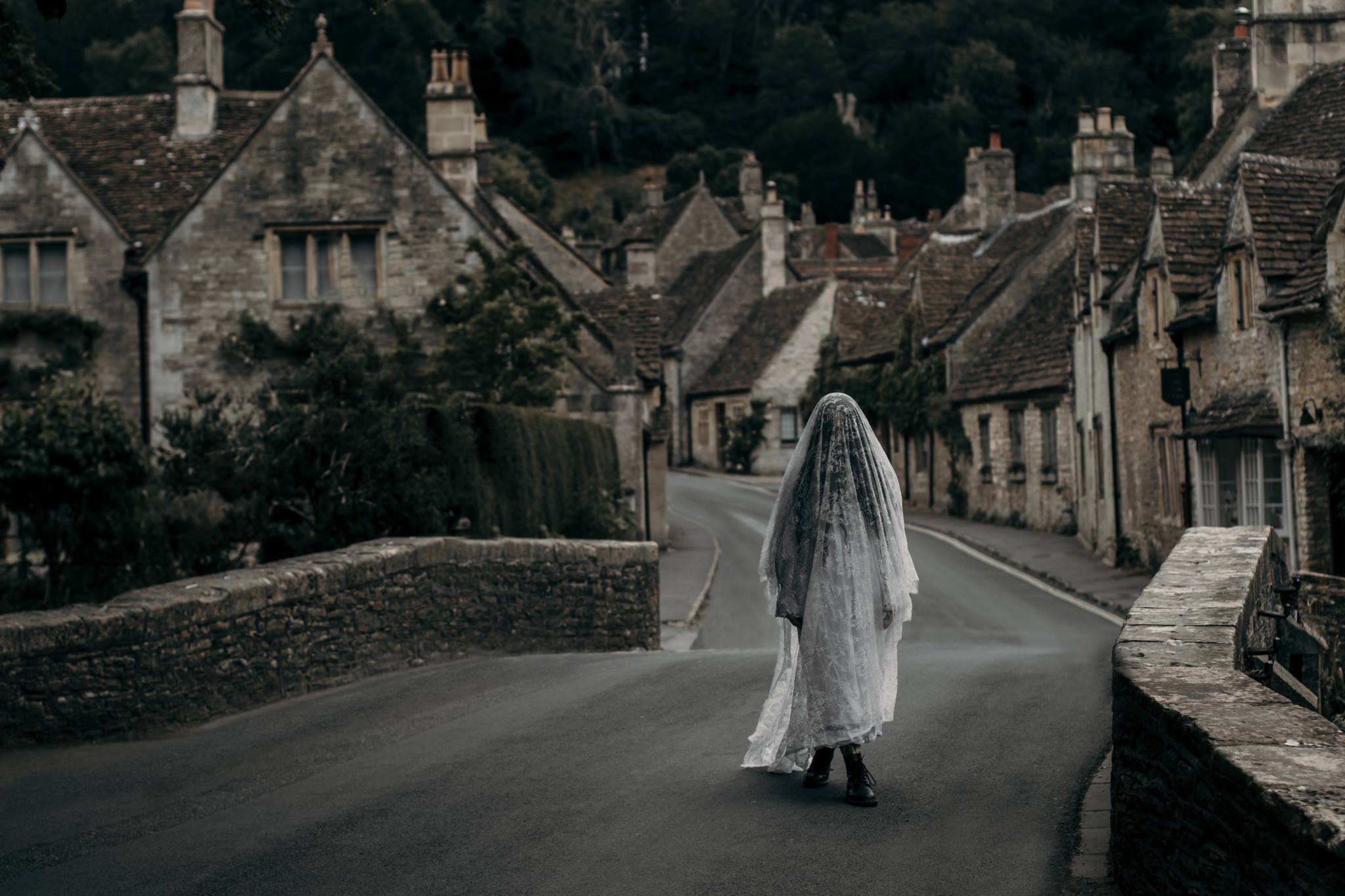 Castle Combe photoshoot - Eerie horror photography of woman in a lace veil standing on the bridge in a medieval English village.