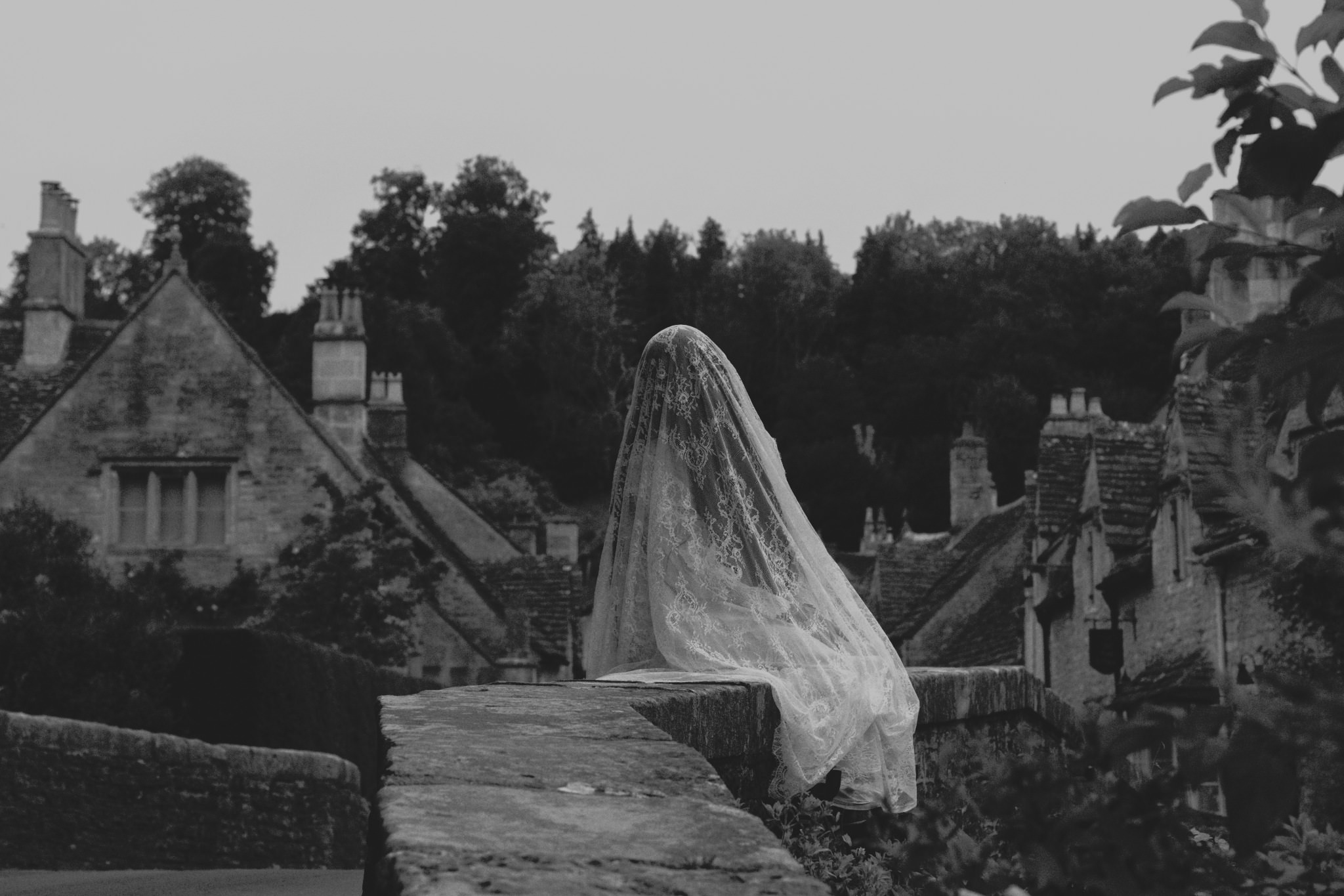 Castle Combe horror photoshoot - Woman in veil with long black hair, sat on the edge of a bridge in an old English village