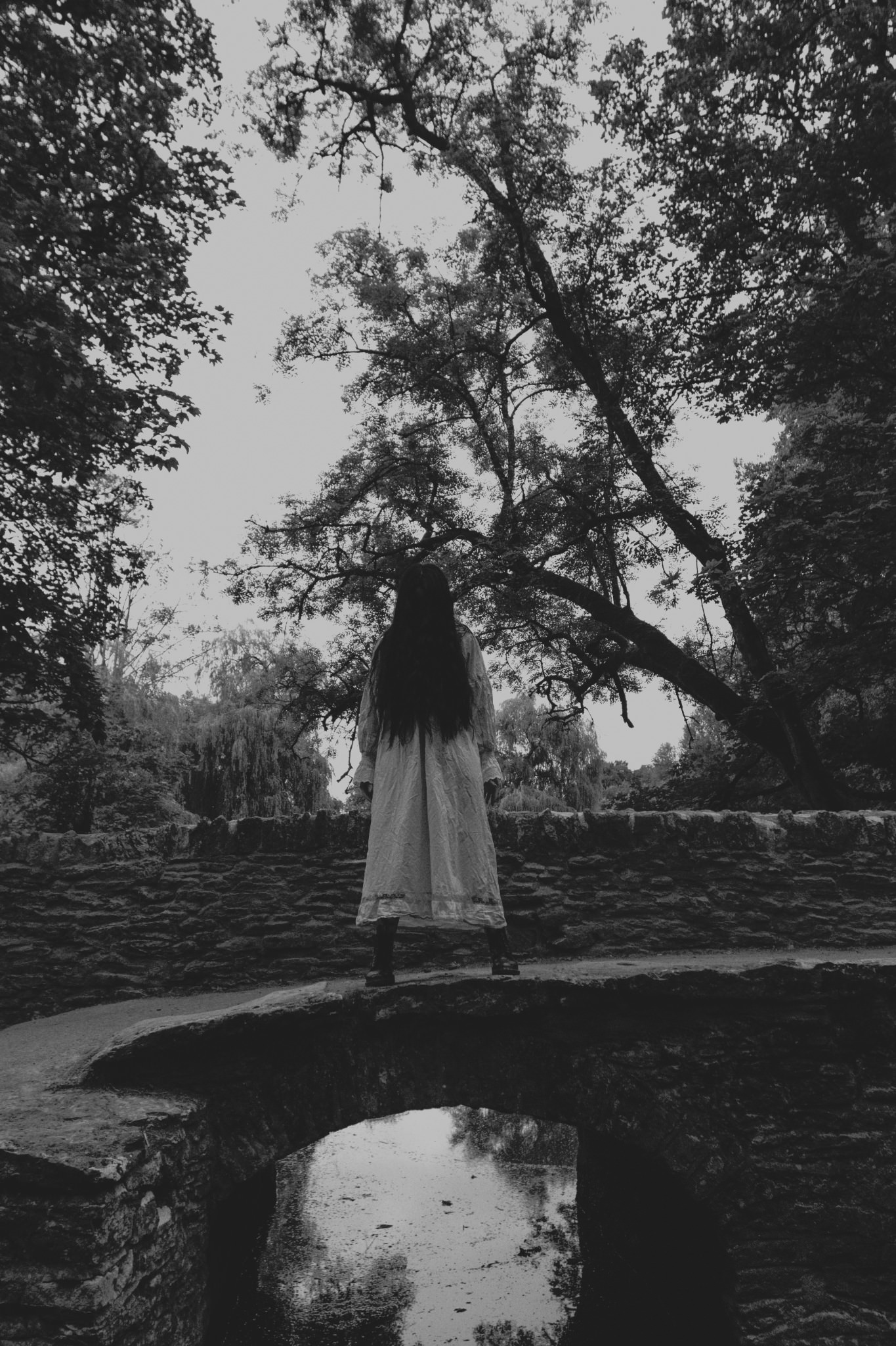 Creepy woman with long black hair and long white nightdress standing on a bridge over a river in Castle Combe photoshoot