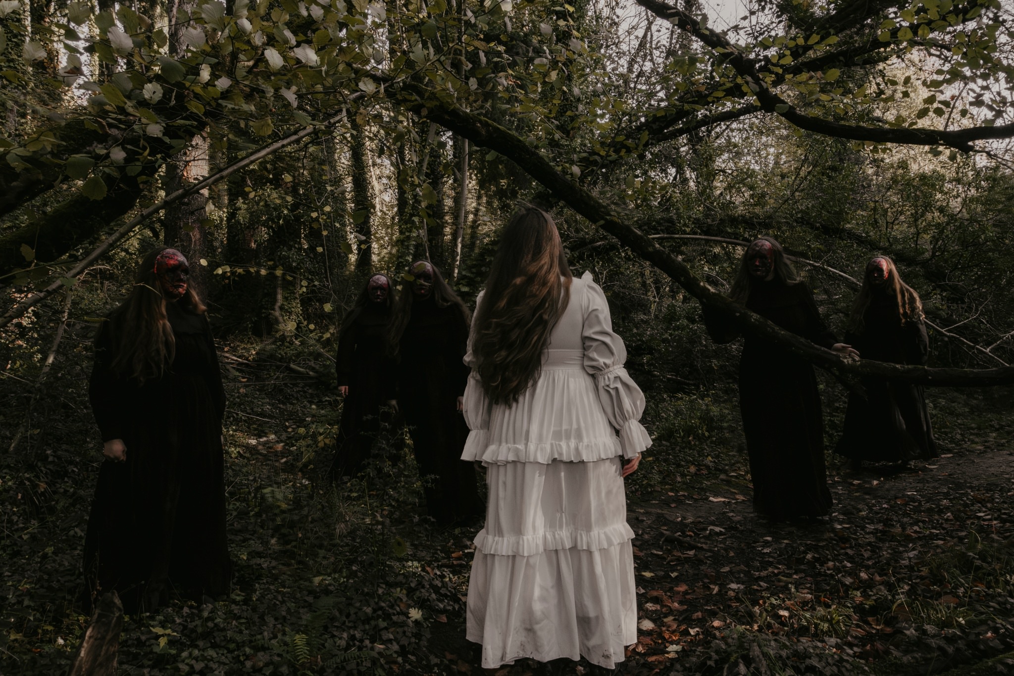 Horror concept photography - woman in white surrounded by dark figures in creepy masks in the woods