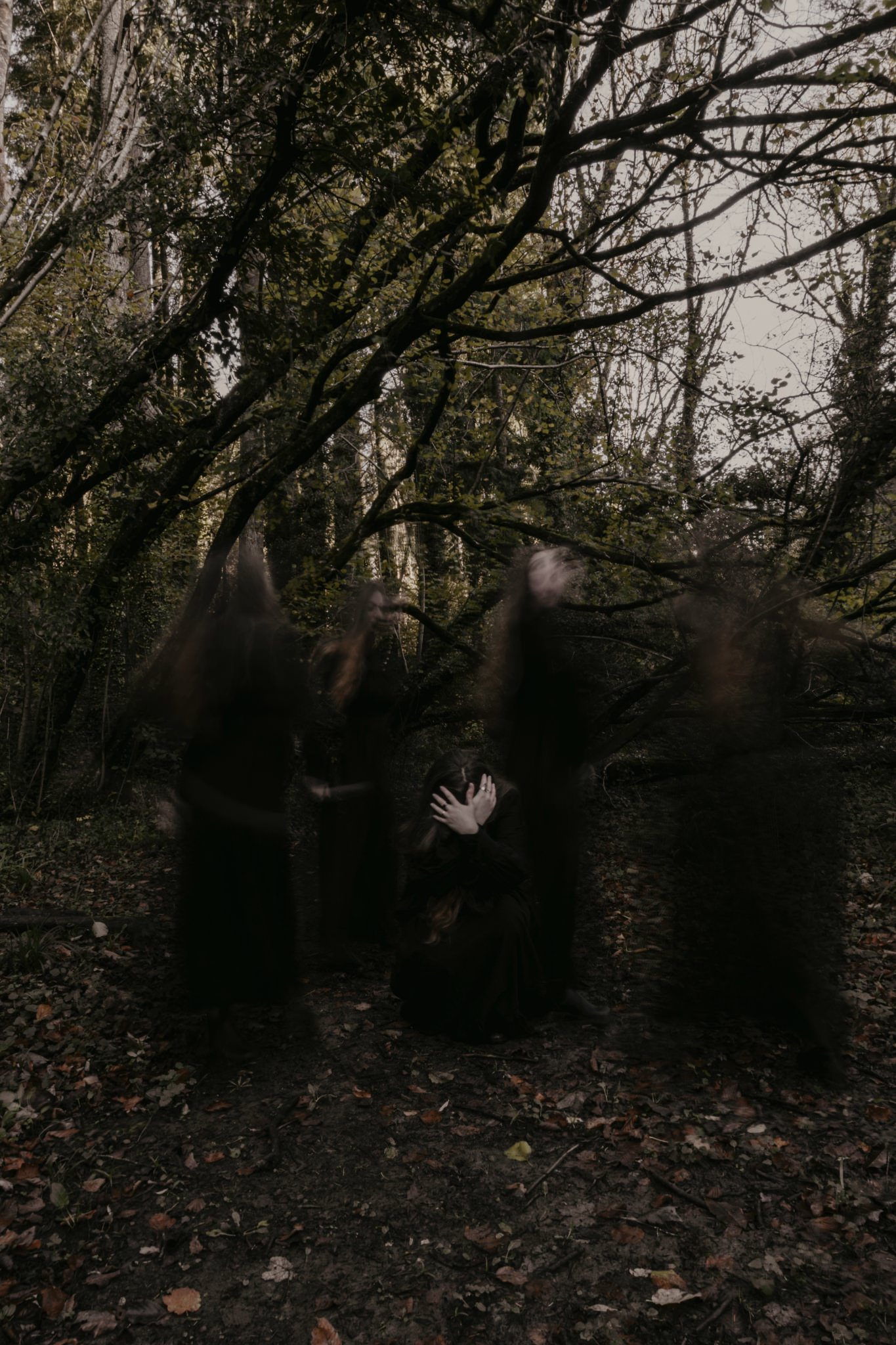 Woman on the ground in a woodland, holding her head and hiding her face, while ghosts and memories surround her.
